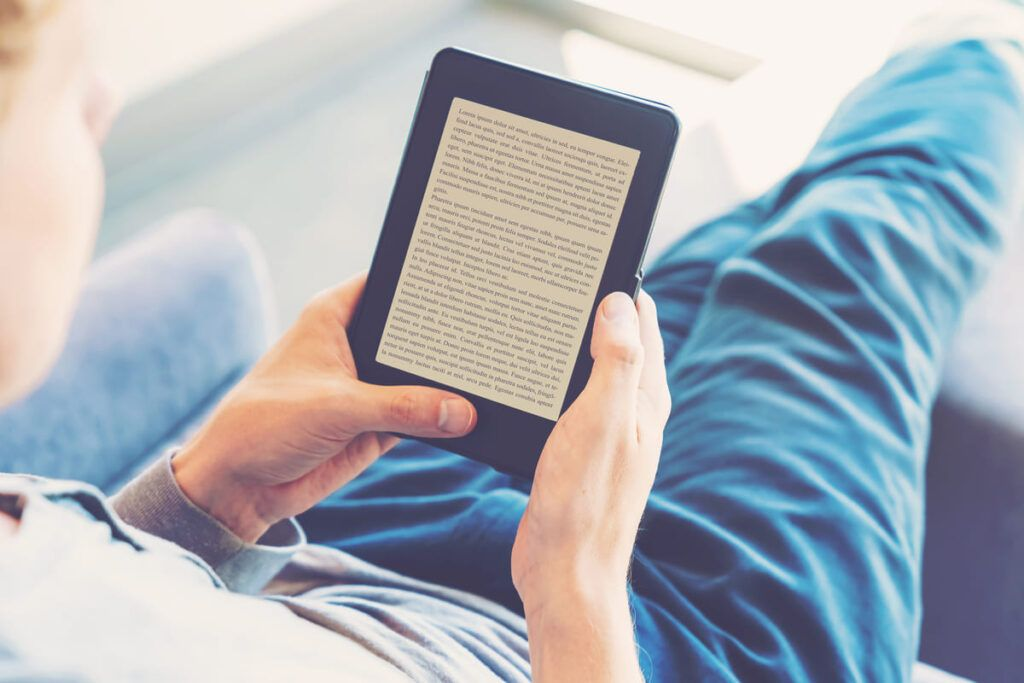 Um homem relaxa em um sofá enquanto lê um e-book em seu dispositivo digital. O consumo de infoprodutos como livros digitais tem se tornado uma alternativa conveniente para o aprendizado.