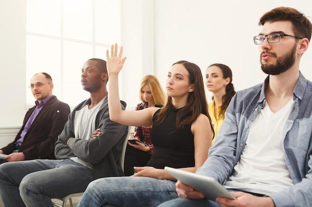 Em um ambiente de aprendizado, uma jovem levanta a mão para participar da discussão. Sua postura e confiança mostram como uma boa dicção é essencial para se expressar com clareza e ser compreendido pelos outros. Ao seu redor, os demais participantes também demonstram interesse, reforçando a importância de uma comunicação eficaz.