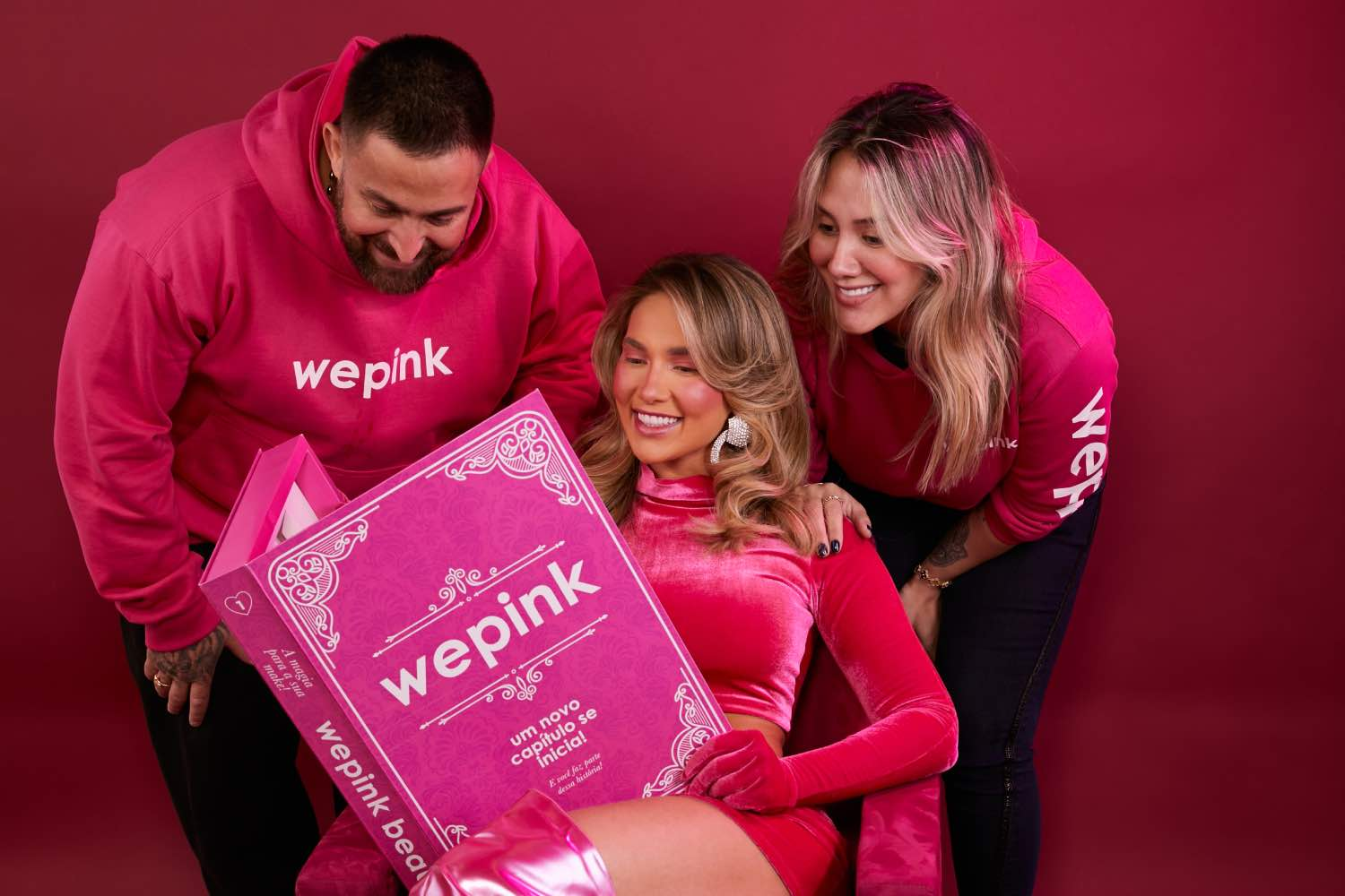 A imagem mostra três pessoas sorrindo, vestindo camisetas rosas da Wepink, enquanto uma delas segura uma grande caixa rosa da marca. A Wepink está se destacando no mercado de cosméticos, e quem deseja criar uma marca com esse tipo de apelo visual pode aprender muito com um curso online sobre branding, design de produtos e estratégias de marketing digital.