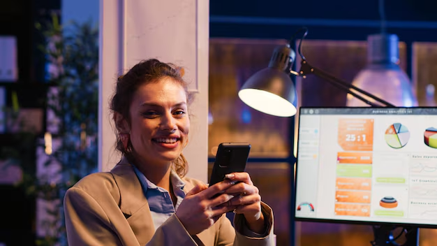 Mulher sorridente segurando o celular em um ambiente corporativo, com gráficos de métricas de social media ao fundo. O sucesso no marketing digital depende de uma estratégia eficaz de mídia social para alcançar mais público.