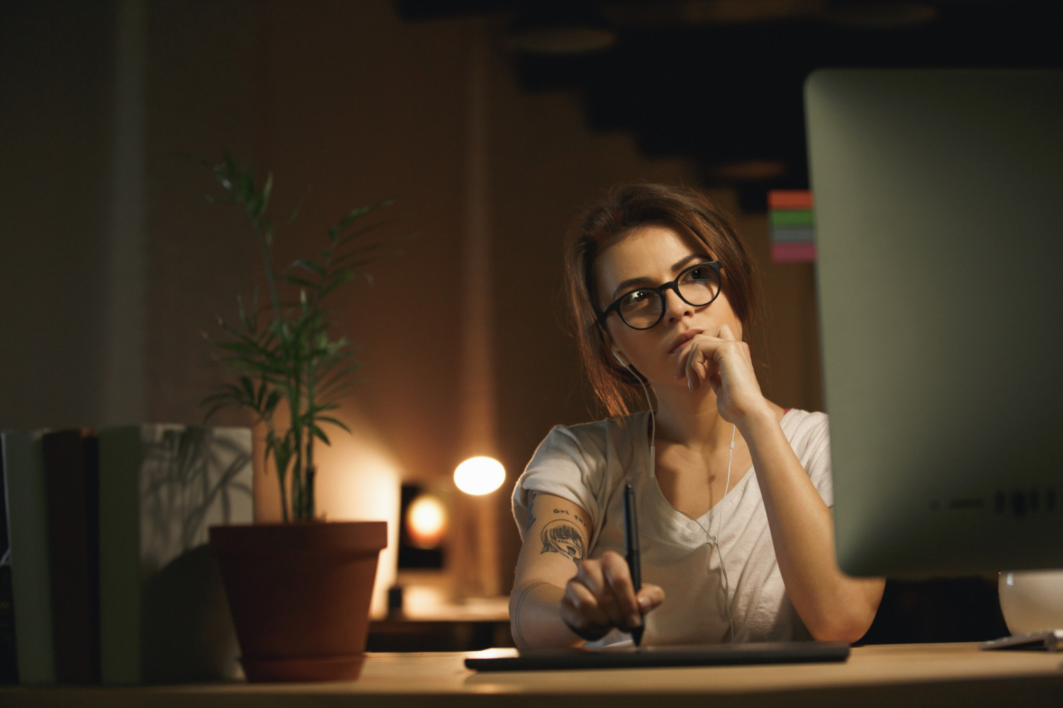 O que é Copy? Os princípios e o que você precisa saber sobre Copy 1 Mulher jovem pensativa trabalhando à noite em frente ao computador, com fones de ouvido e caneta digital na mão, refletindo sobre a criação de uma copy envolvente para conteúdo digital.