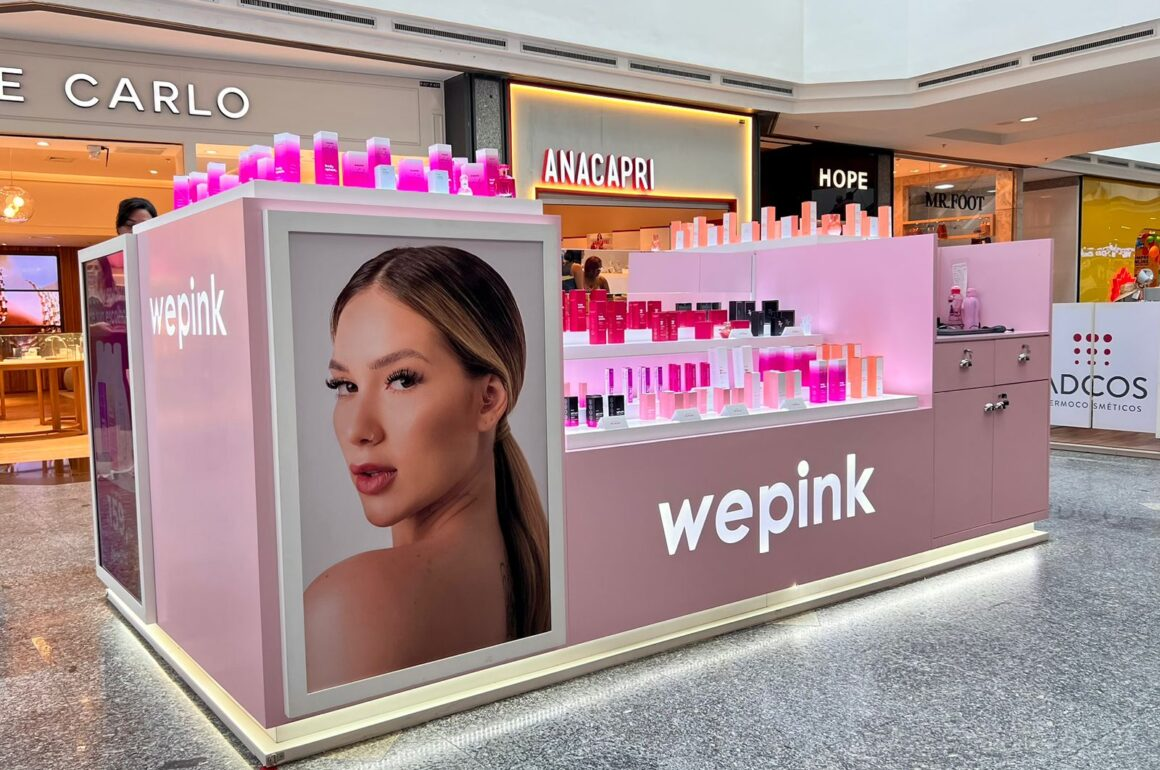 Foto de um quiosque da Wepink em um shopping, com produtos cosméticos nas cores rosa e preto. O quiosque é decorado com iluminação brilhante e uma grande imagem de uma mulher na lateral. Se você tem interesse em aprender como lançar uma marca como a Wepink, um curso online de marketing ou empreendedorismo pode ser o próximo passo para o sucesso.