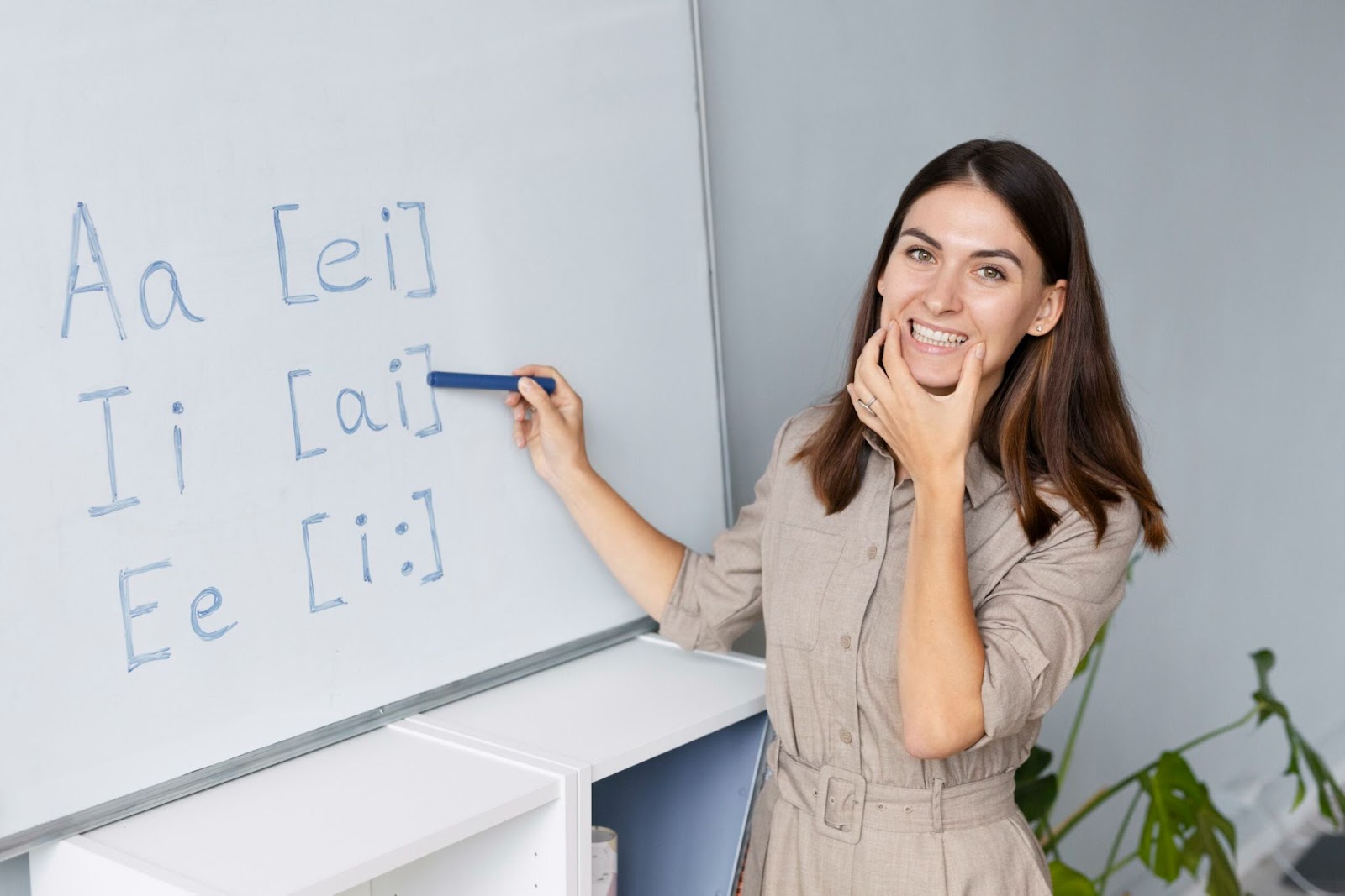 Como Melhorar sua Dicção em Menos de 1 Minuto 4 Uma mulher sorridente, professora de idiomas, aponta para um quadro branco onde estão escritos as letras 'A', 'I', 'E' e suas respectivas pronúncias fonéticas. A imagem simboliza o aprendizado da dicção e da pronúncia de novas palavras e fonemas, enfatizando a importância da clareza na fala.