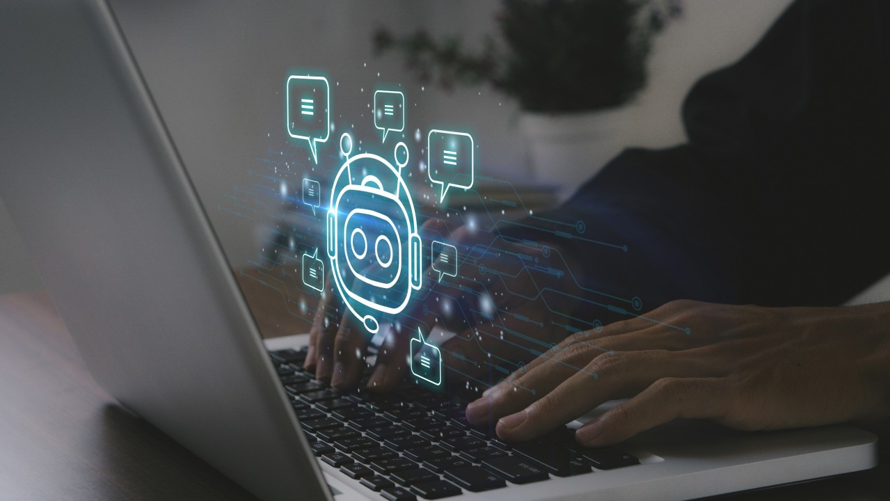 Como Usar a Inteligência Artificial? 7 Dicas Valiosas Para o Seu Negócio 9 Uma imagem de perto das mãos de uma pessoa digitando em um laptop, com um ícone de chatbot luminoso e holográfico projetado acima do teclado. A imagem, com foco em Inteligência Artificial, ilustra a experiência do usuário com assistentes virtuais ou plataformas de conversação baseadas em IA.
