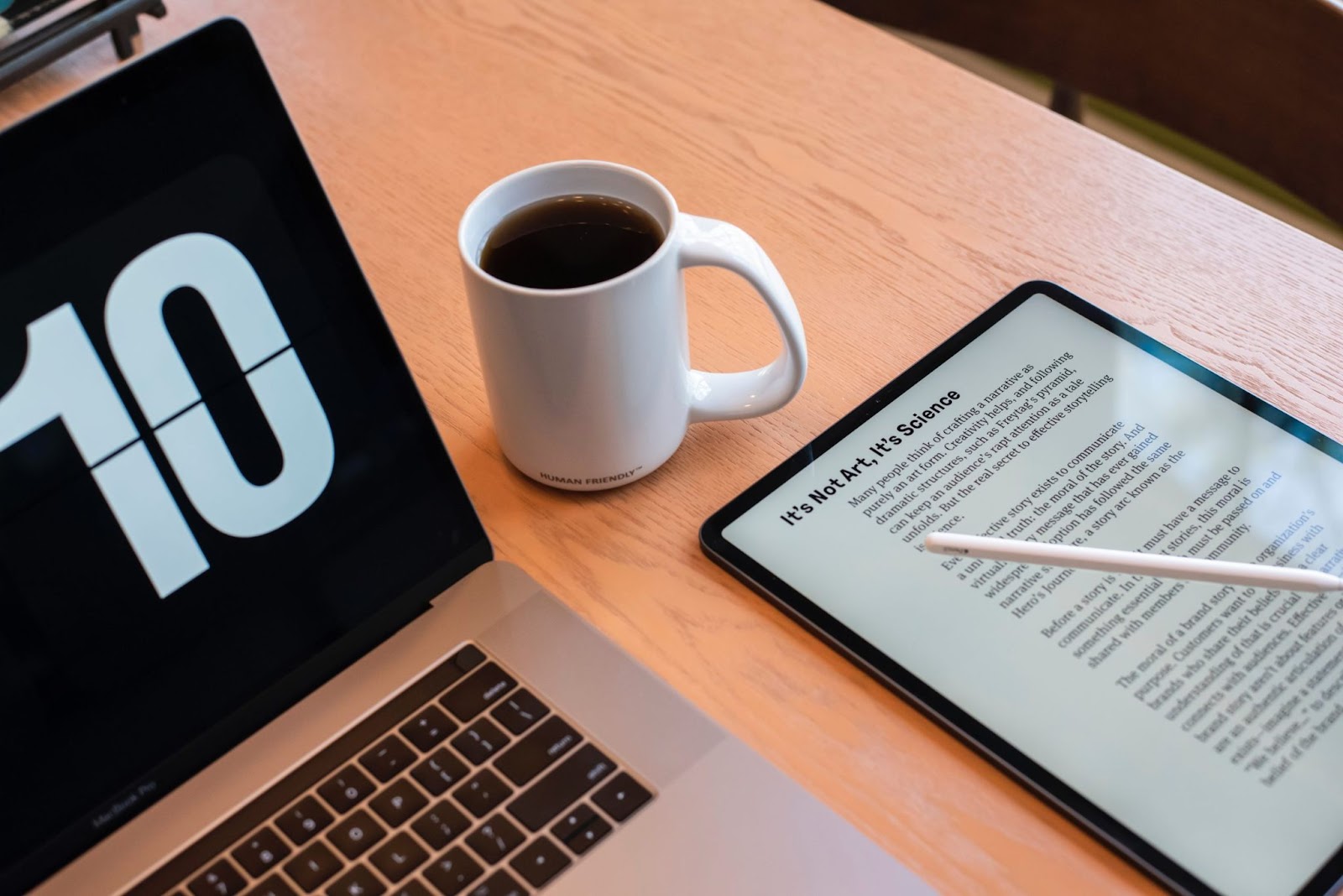 Um tablet com um artigo aberto, um notebook e uma xícara de café sobre uma mesa de madeira. O título do artigo no tablet é 'It's Not Art, It's Science', e as ferramentas de trabalho representam a rotina de um profissional que consome e cria produtos digitais.