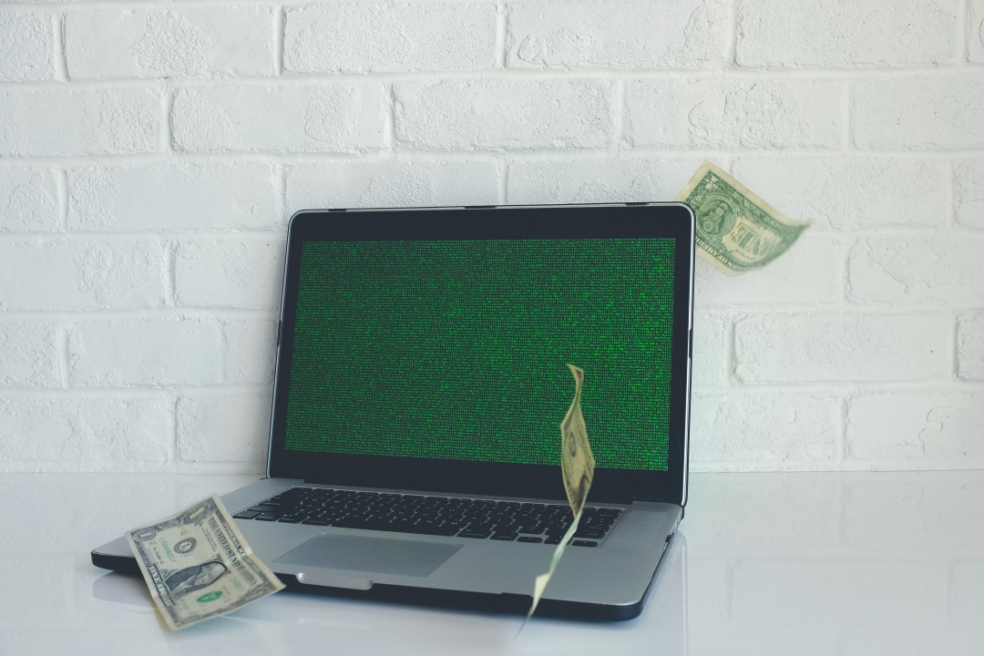 Laptop sobre uma mesa branca com notas de um dólar voando ao redor. A tela do computador exibe códigos binários ou de programação em cor verde. A imagem simboliza como o tráfego na internet se converte em dinheiro ou a gestão de tráfego pago (investimento versus retorno).