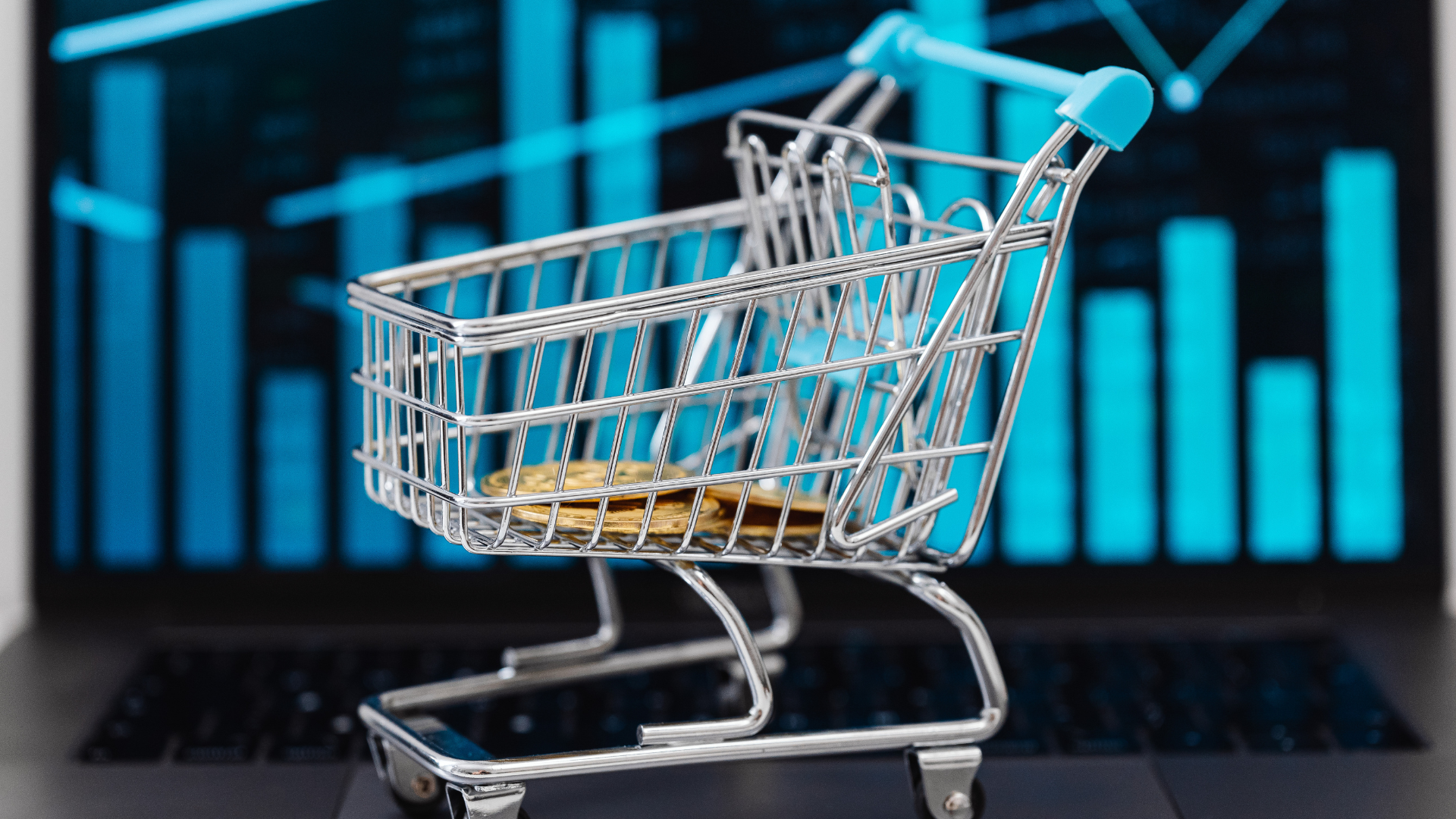 Uma Aula de Vendas que Vale Milhões – Script Bônus no Final 5 Um mini carrinho de compras de metal com detalhes em azul claro está posicionado sobre o teclado de um laptop. Ao fundo, a tela do computador exibe gráficos de barras e indicadores financeiros brilhantes em tom azulado, ilustrando o monitoramento de métricas e performance de vendas digitais.