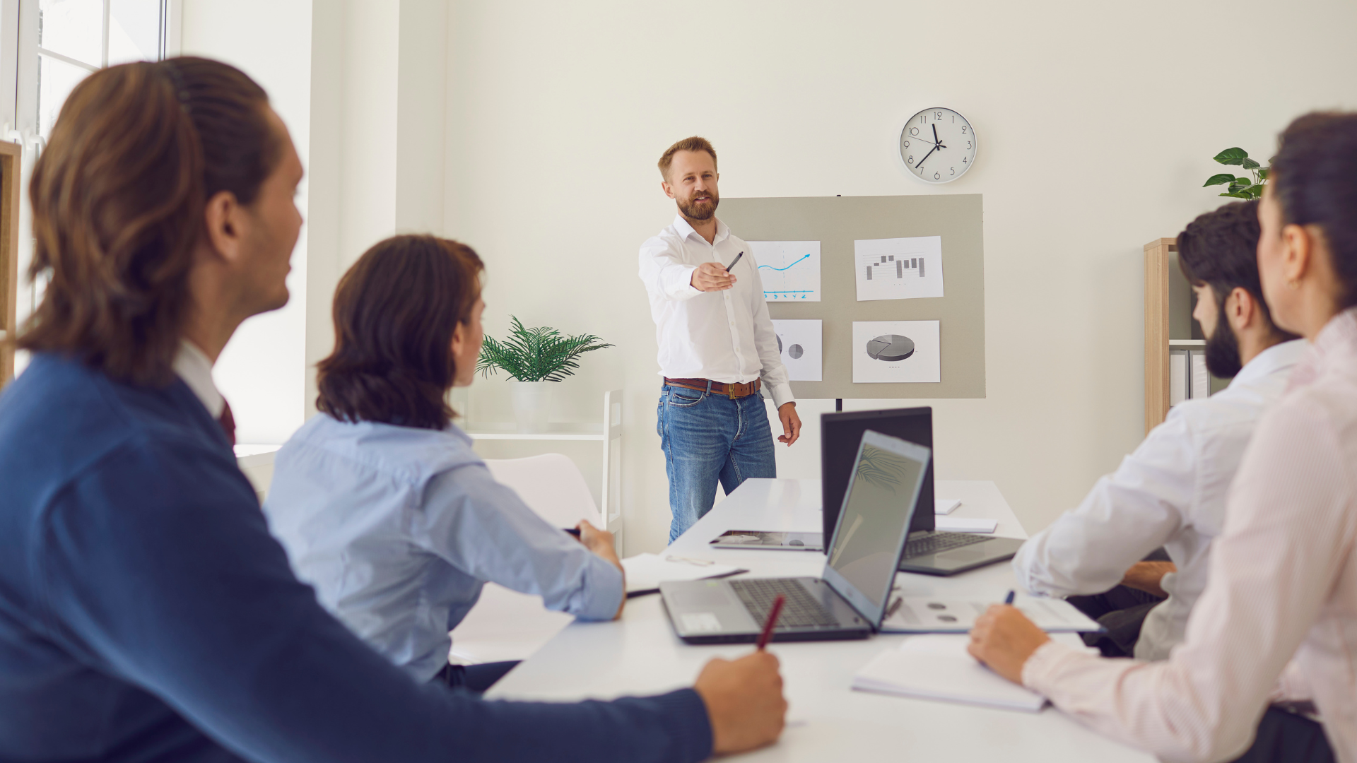 Uma Aula de Vendas que Vale Milhões – Script Bônus no Final 1 Um homem de camisa branca e calça jeans apresenta gráficos de desempenho em um quadro para uma equipe corporativa sentada ao redor de uma mesa de reuniões com laptops. A cena ilustra um treinamento ou alinhamento estratégico com foco em impulsionar as Vendas da empresa.